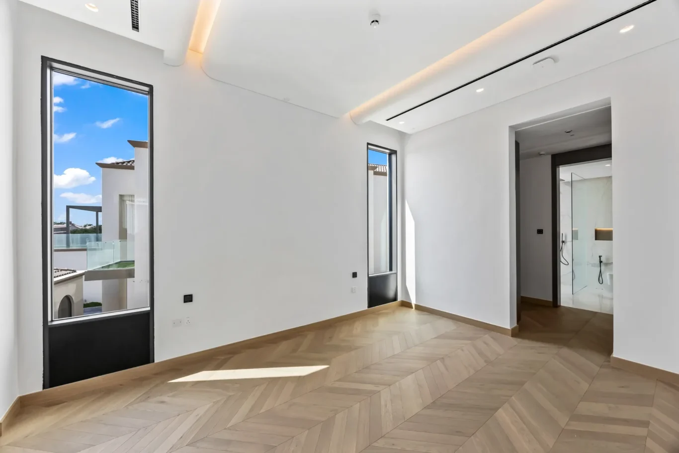 Bright, modern room with white walls, large windows, and a herringbone wood floor; open doorway leads to a glass-walled bathroom.