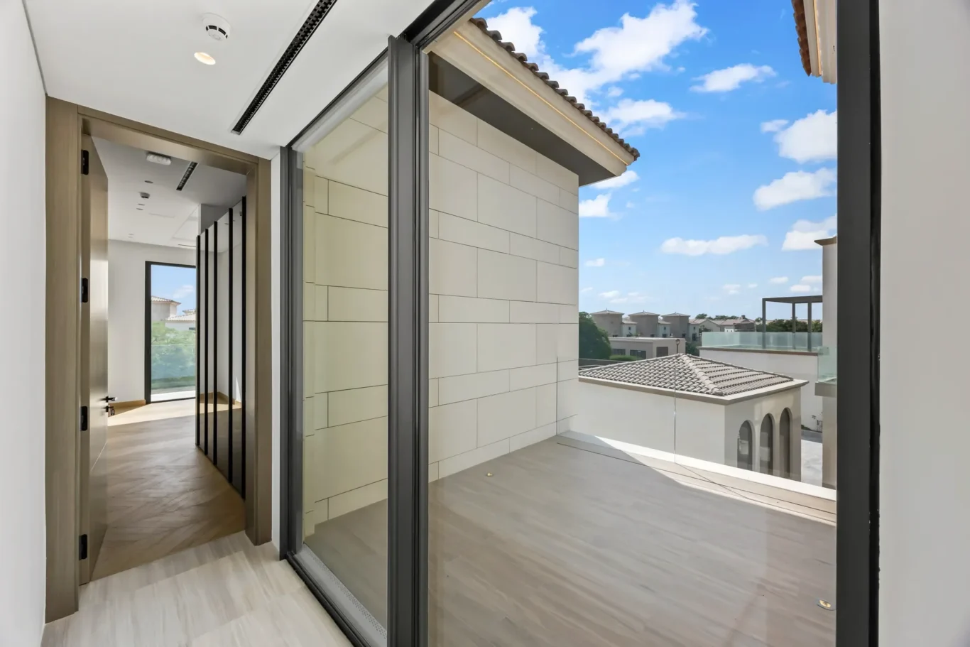 Bright hallway with large black-framed sliding glass doors that open to a sunny balcony and neighboring buildings under a blue sky.