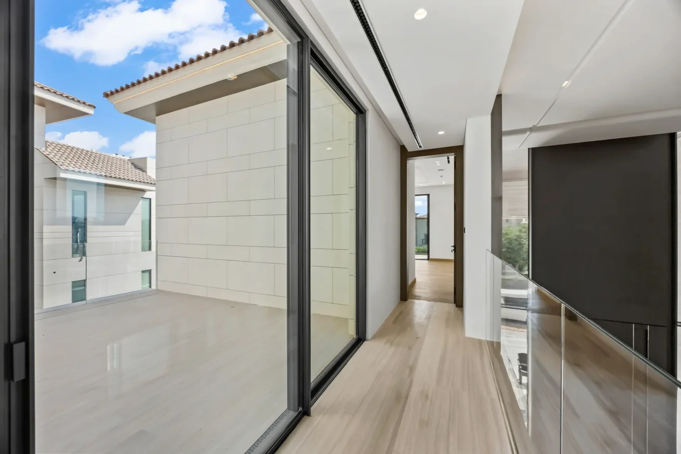 Bright modern hallway with floor-to-ceiling glass doors opening to a balcony and a clear blue sky outside.