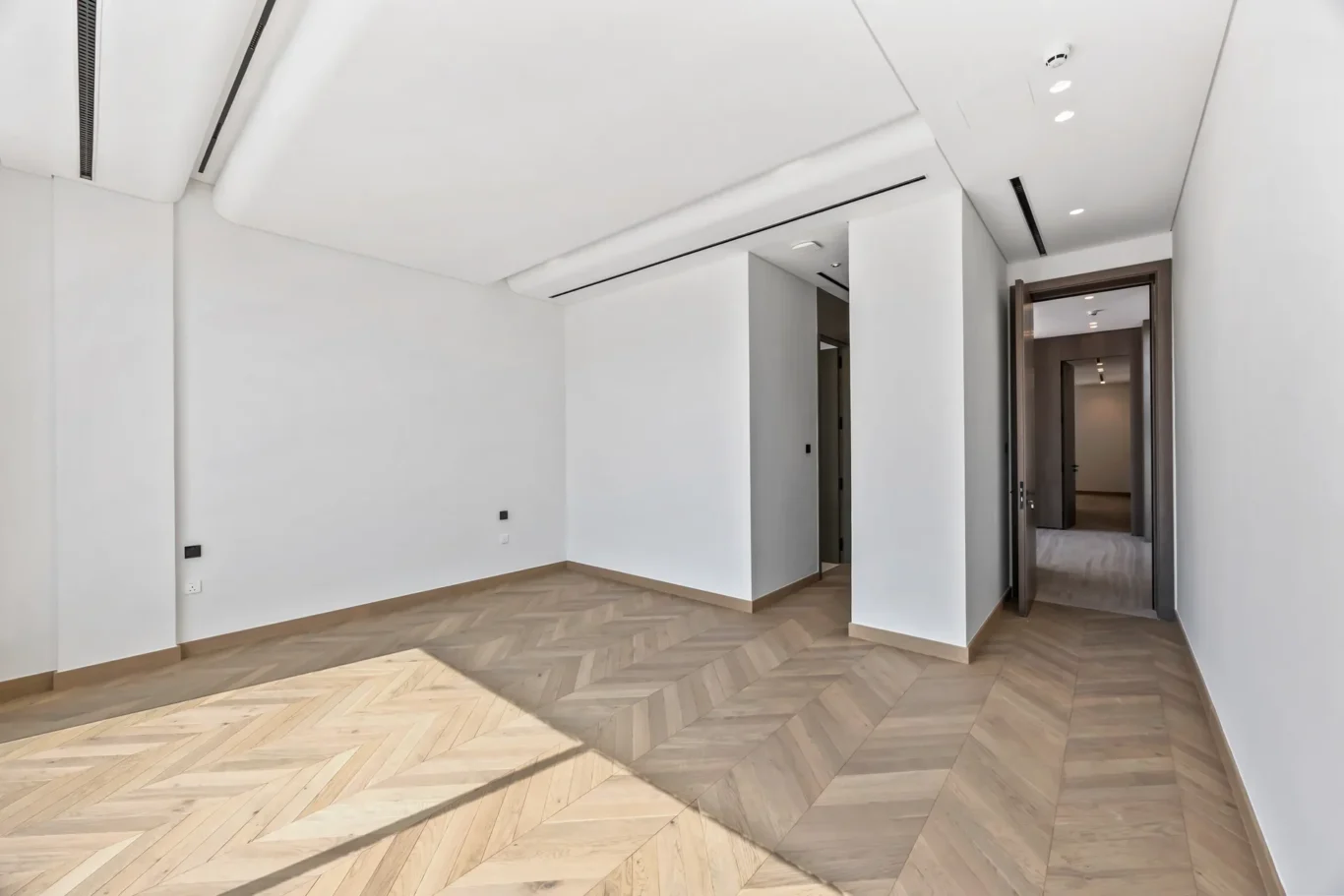 Bright empty modern room with white walls, herringbone wood floor, and a doorway leading to a hall beyond; ceiling vents visible above.