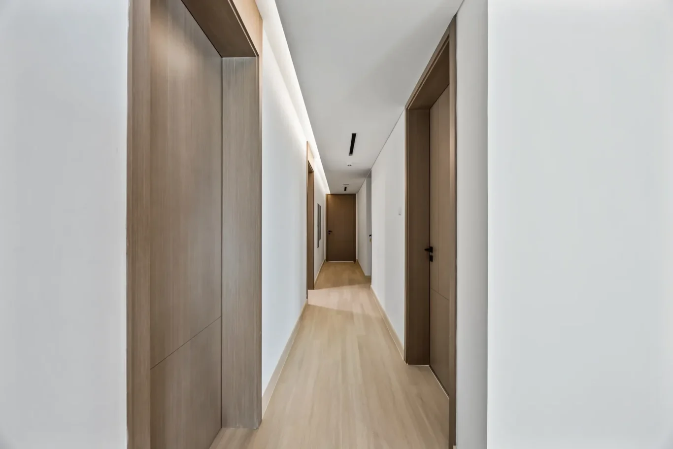 Long modern hallway with light wood flooring and brown wooden doors along white walls, viewed from the foreground toward a door at the end.