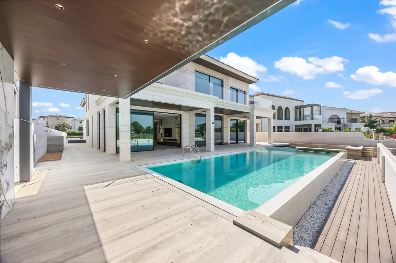 Modern two-story white villa with a rectangular turquoise pool, glass doors, and a wooden deck under a shaded ceiling patio.
