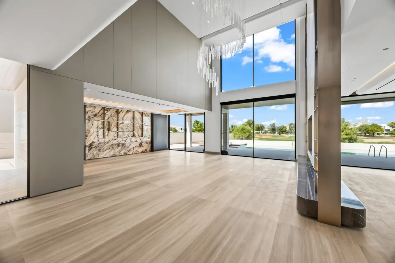 Open modern lobby with tall windows looking out to a blue sky, a marble wall, and a hanging crystal chandelier.