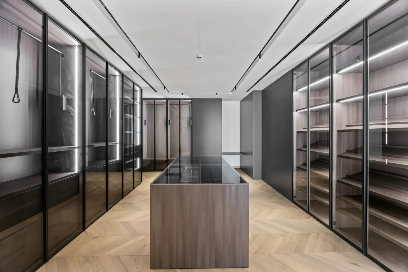 Modern walk-in closet or showroom with glass-front cabinets along both walls and a central wood-toned island with a glass top