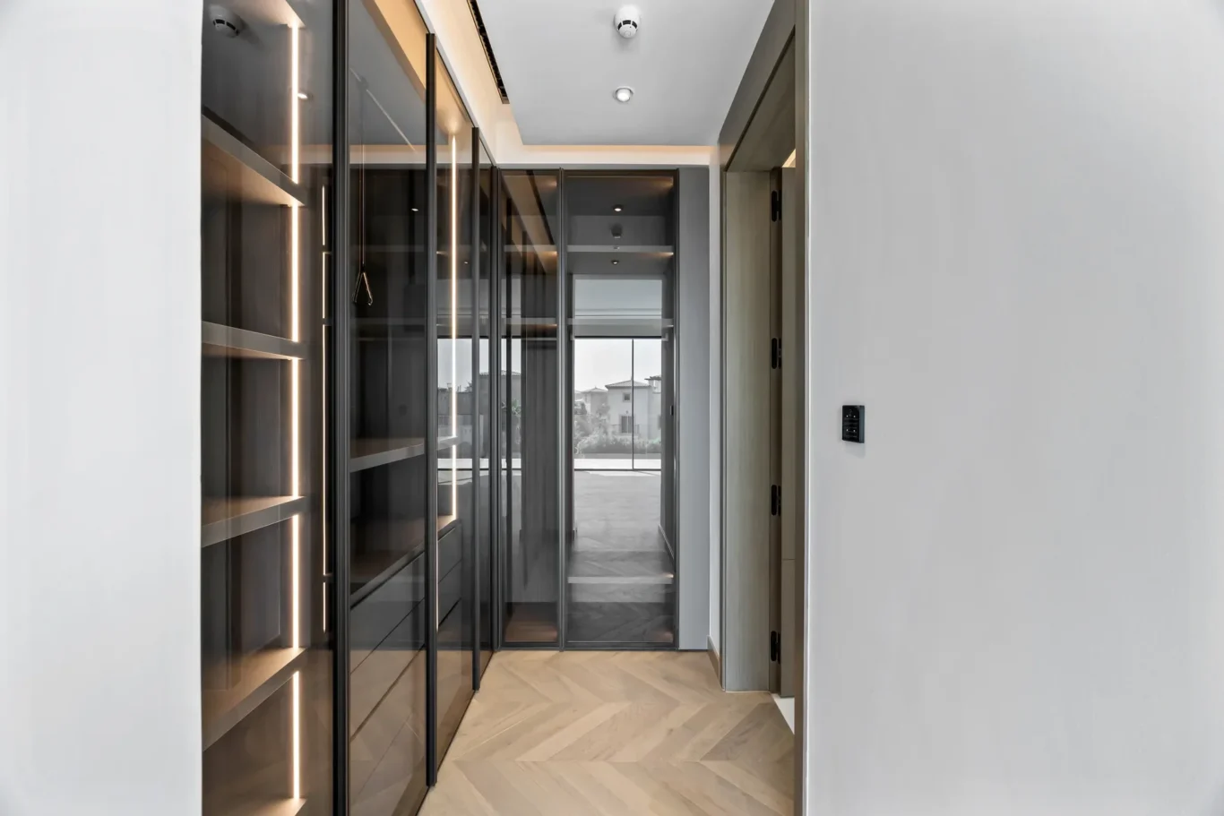 Contemporary hallway with dark glass-front cabinets and built-in lighting, leading to a bright window at the end.