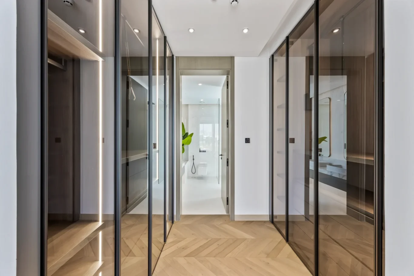 Modern glass-fronted walk-in closet hallway with glossy doors and warm wood flooring leading to a bright bathroom.