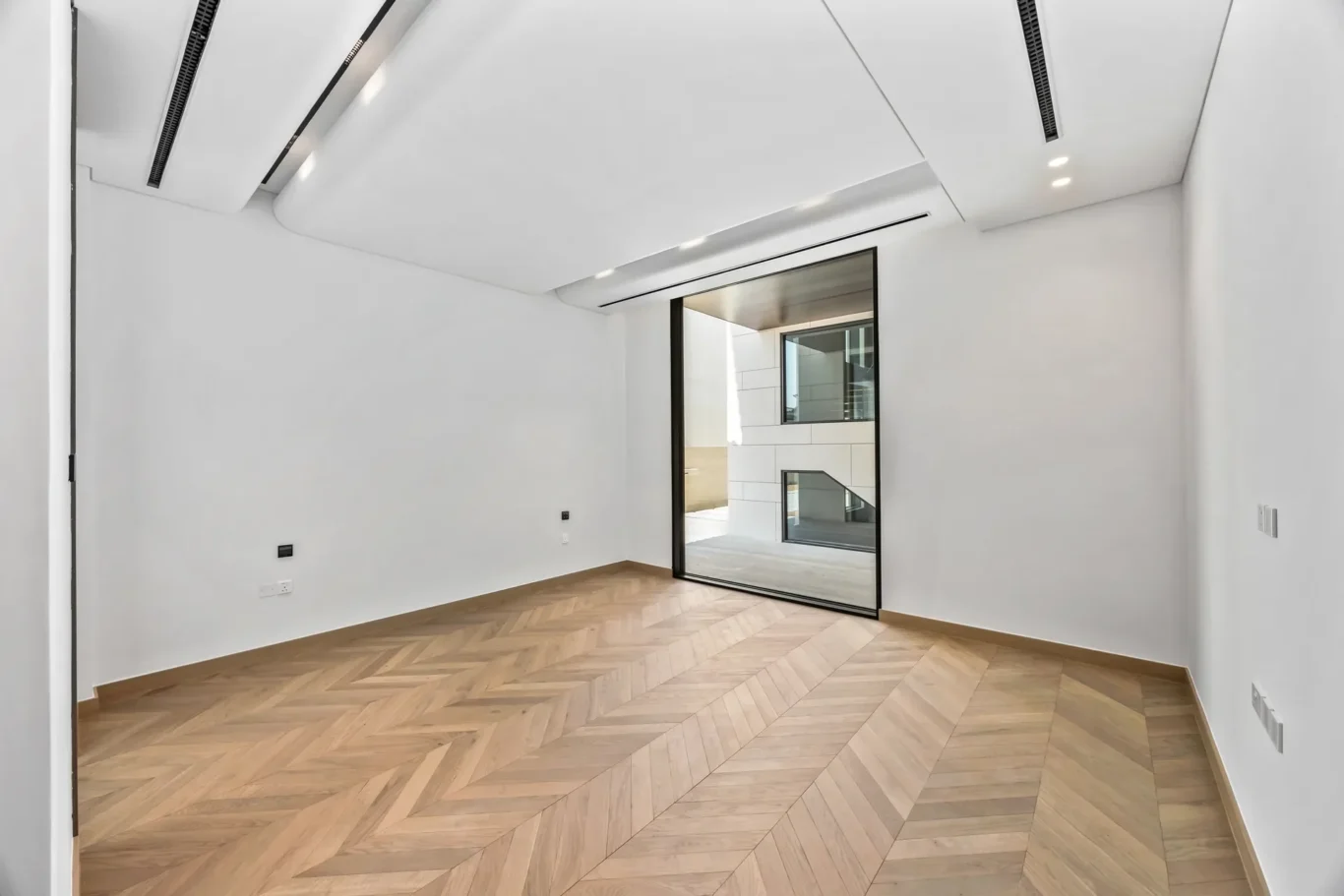 Bright empty room with white walls, herringbone wood floor, and a large sliding glass door leading to an adjacent modern area outside.