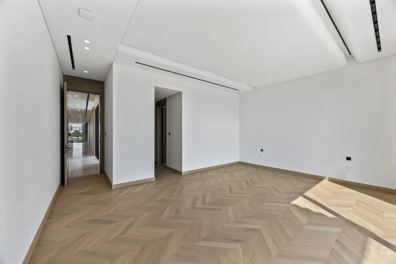Empty modern room with white walls and a herringbone wood floor, hallway visible at the far end with natural light.