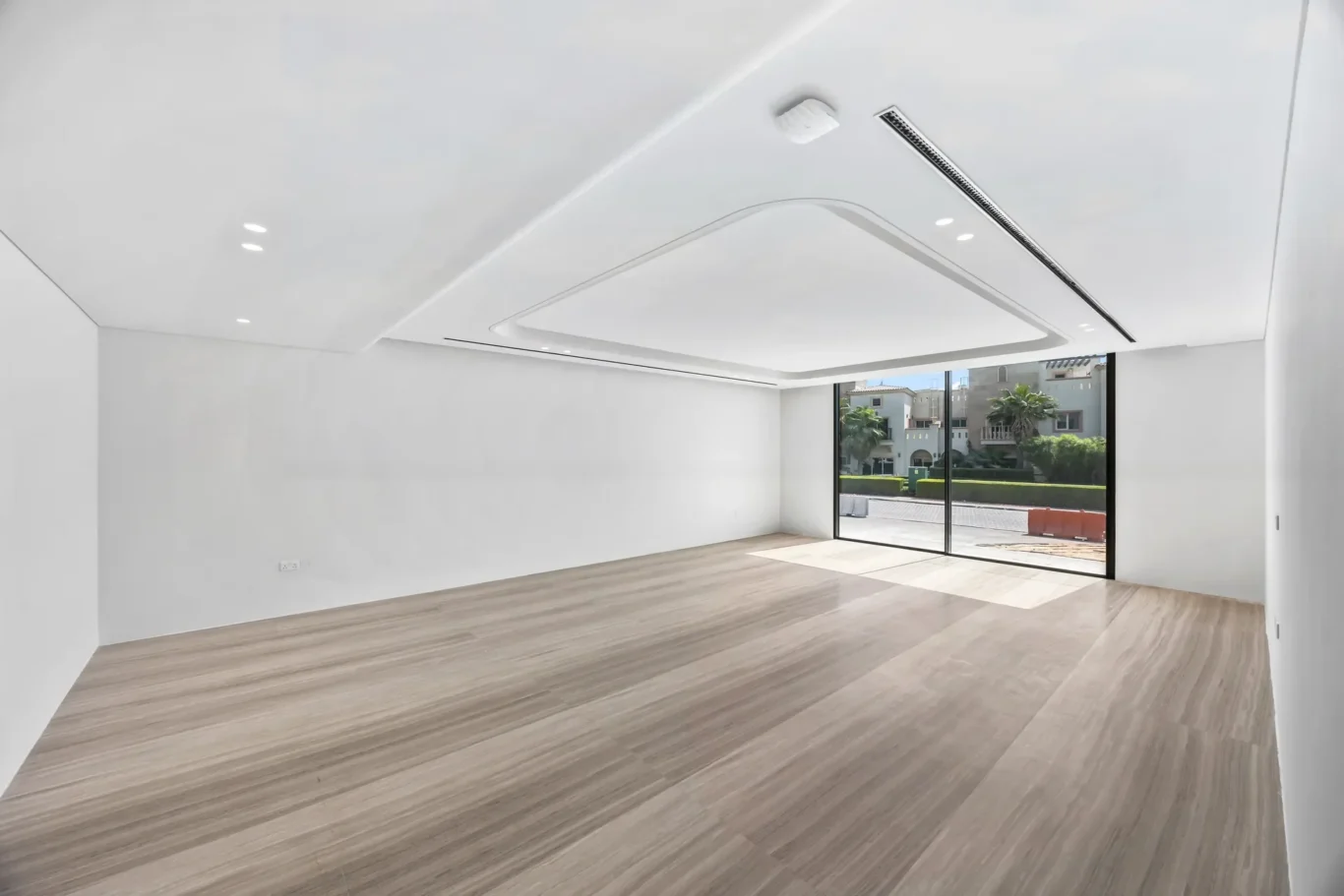 Empty white room with wood floors and a large sliding glass door showing a street with palm trees outside.