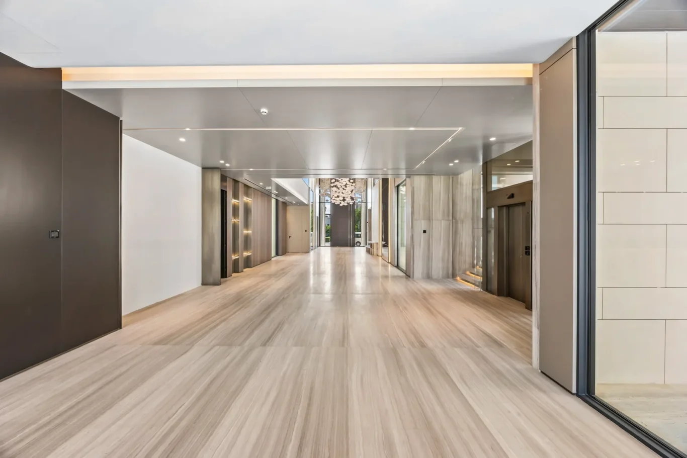 Wide, modern office lobby with elevator banks on both sides, light wood floors, and a decorative chandelier visible at the far end