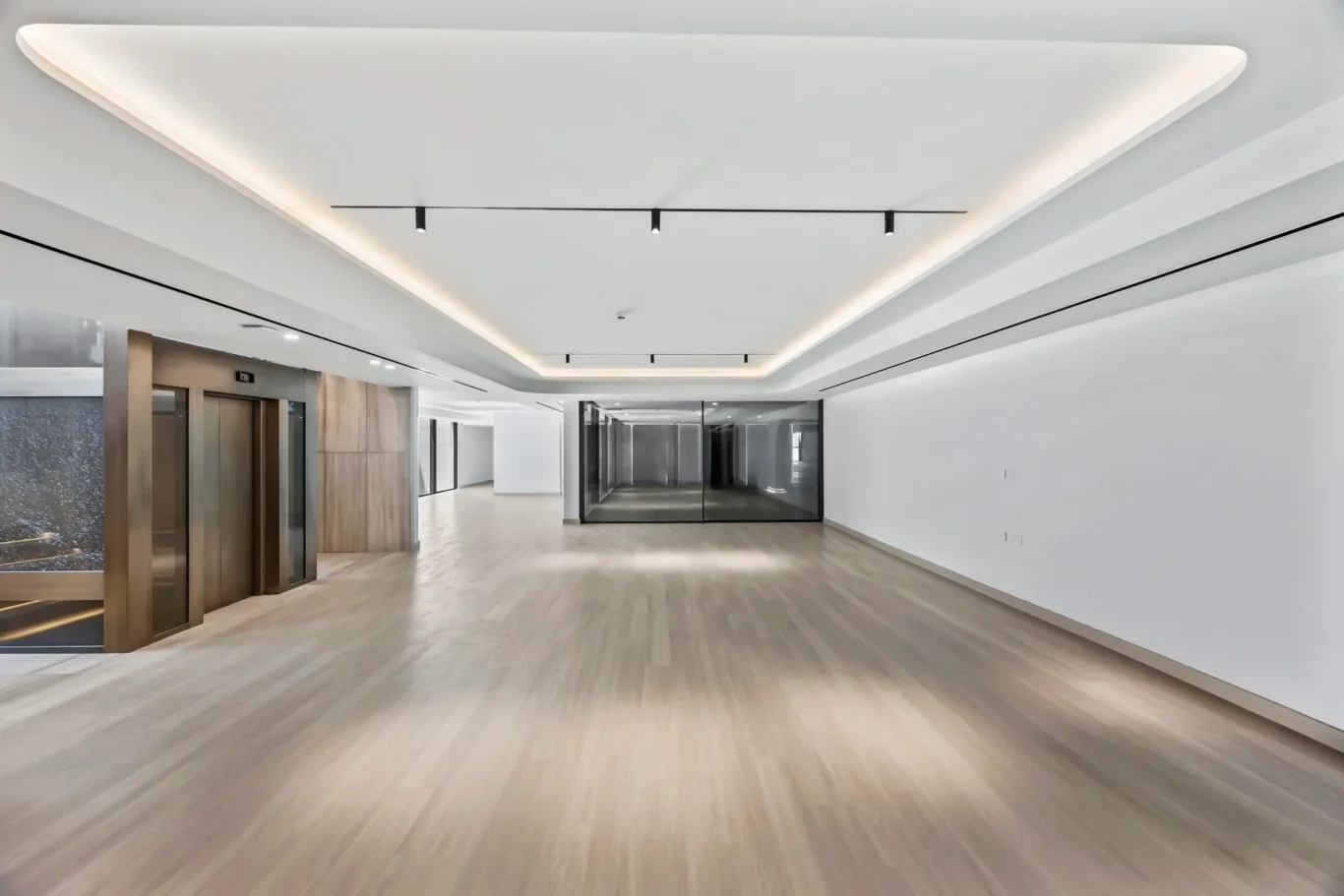 Modern lobby hallway with an elevator on the left, glass partitions, and warm wood flooring under recessed ceiling lights.