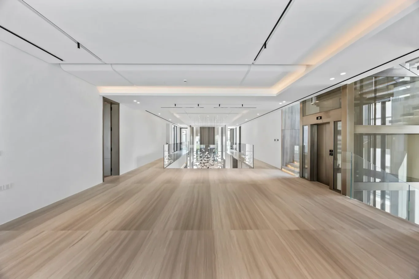 Wide-angle view of a bright, modern office lobby with white walls, light wood floors, glass railings, and elevators on the right along a long corridor.