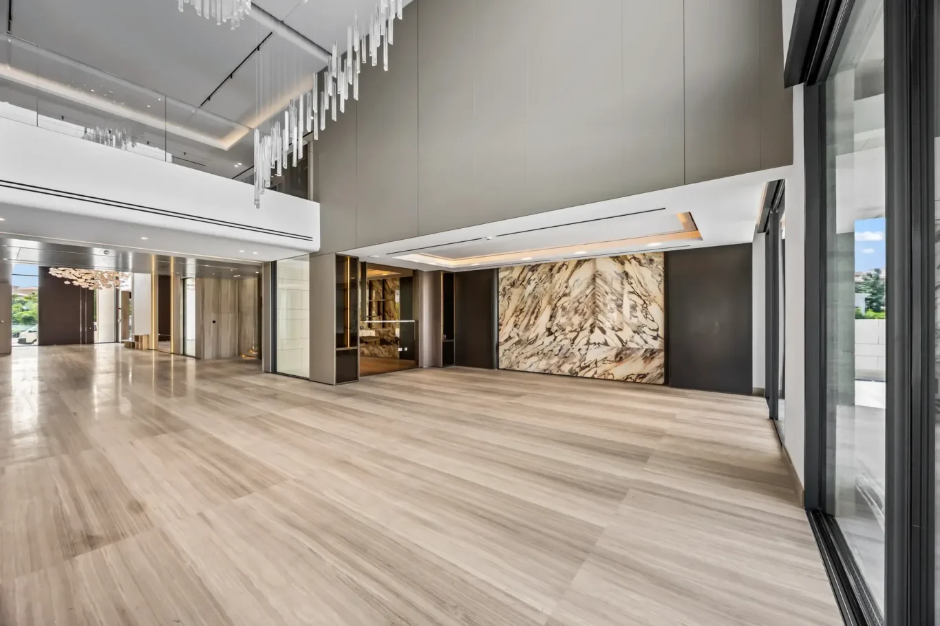 Spacious modern lobby with light wood flooring, a marble feature wall, glass doors, and a hanging crystal chandelier for an upscale reception area