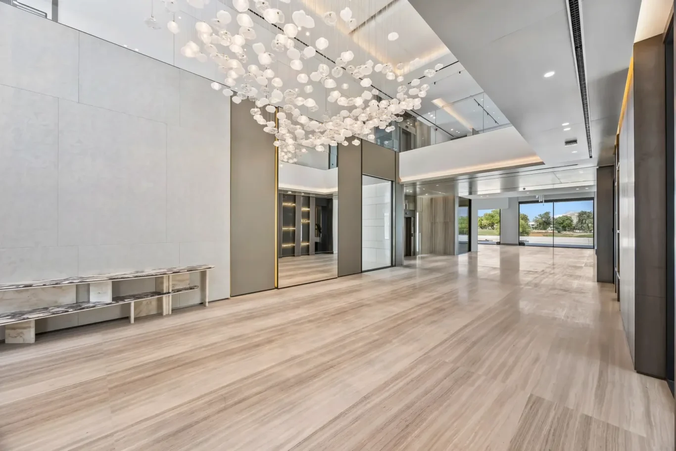 Spacious modern lobby with striped marble floor, glass walls, and a hanging cluster of decorative glass globes above a long marble bench on the left.