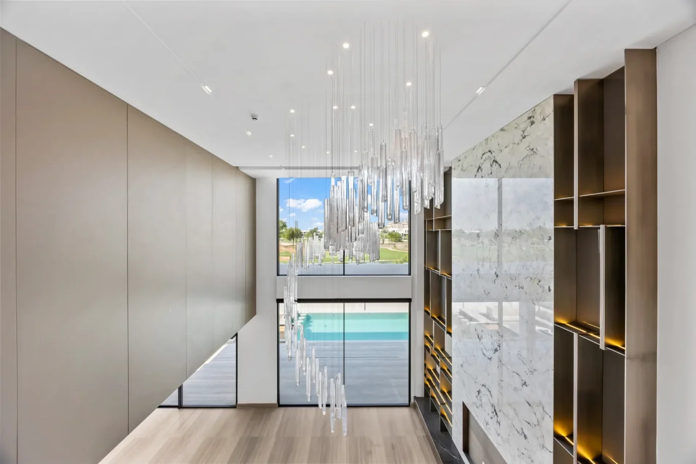 Modern foyer with a cascading crystal chandelier, beige wall panels, and marble shelving along the right, overlooking a pool outside.