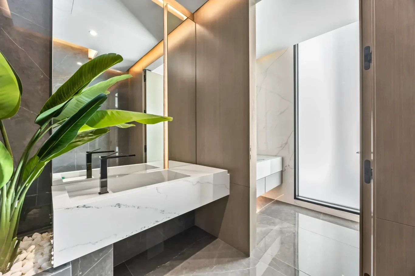Modern bathroom vanity with white marble countertop, black square faucets, wood paneling, and a tall green plant beside a mirror.