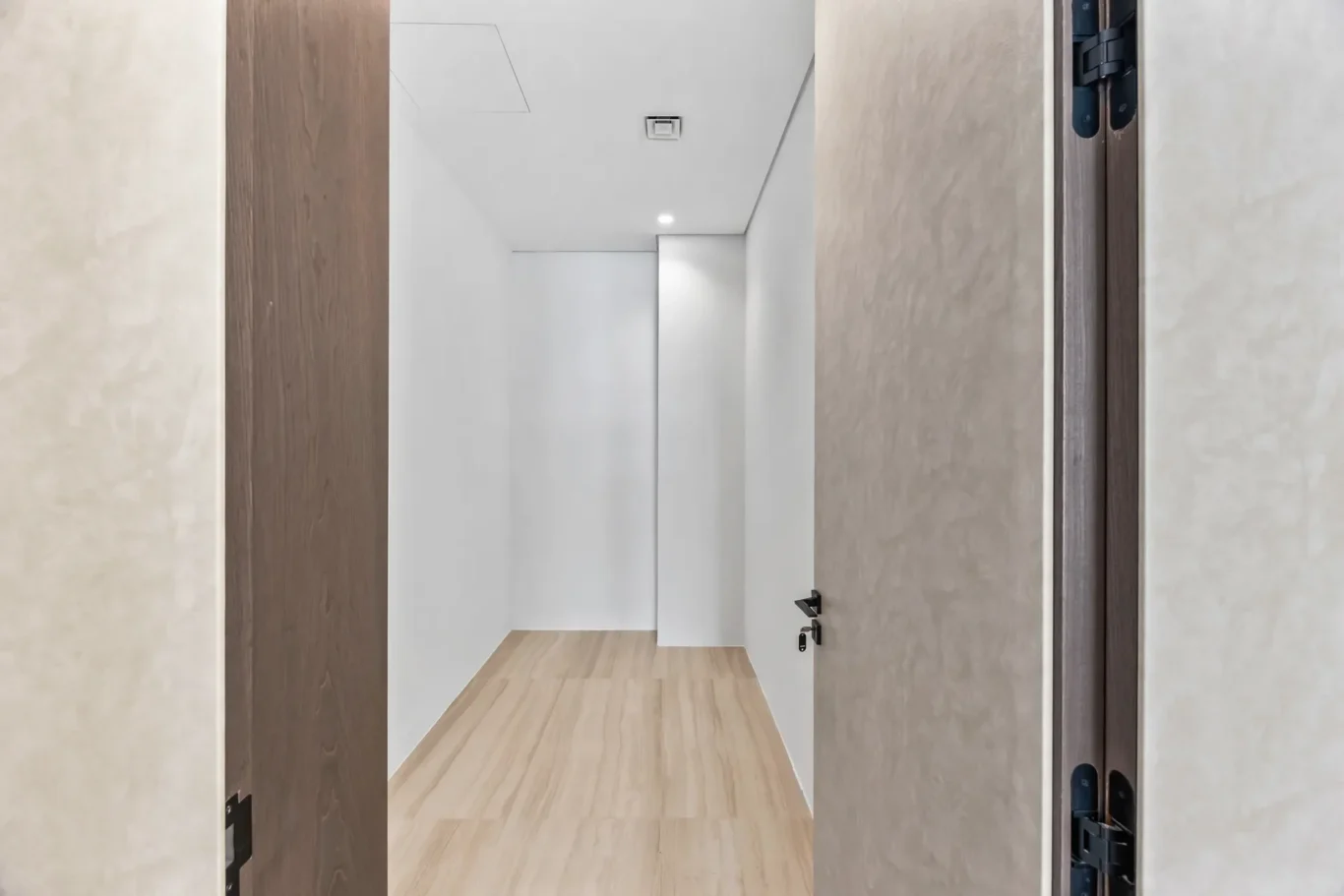 View down a narrow hallway with white walls, light wood floor, and open white doors on both sides.