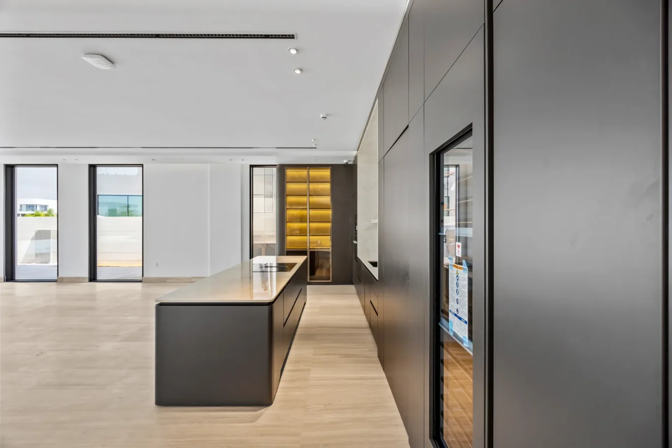 Modern kitchen showroom with a large island, dark gray cabinets, and glass doors along a bright open space.