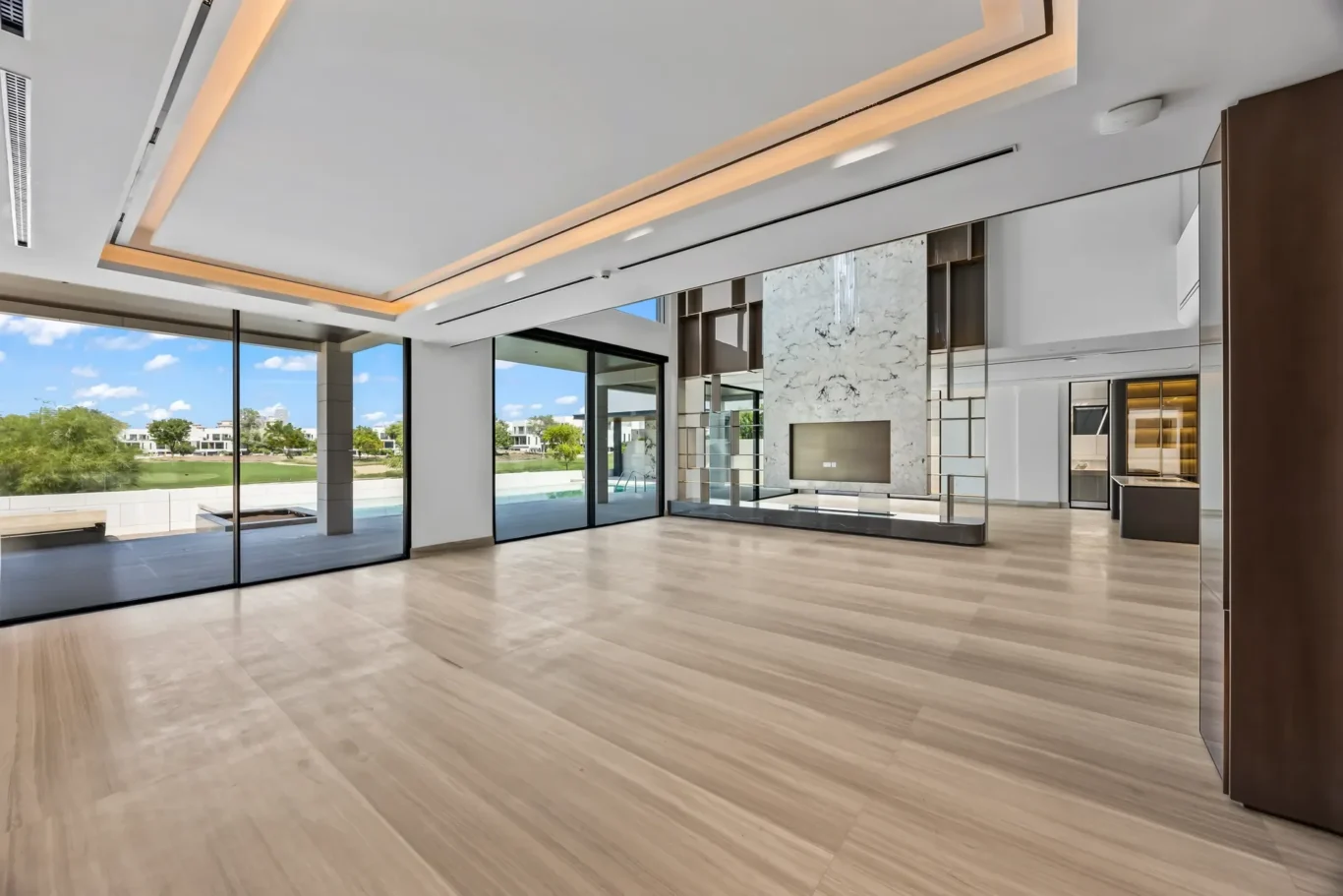 Spacious modern lobby with large glass doors, light wood floors, and marble fireplace centerpiece; outdoor greenery visible through windows.