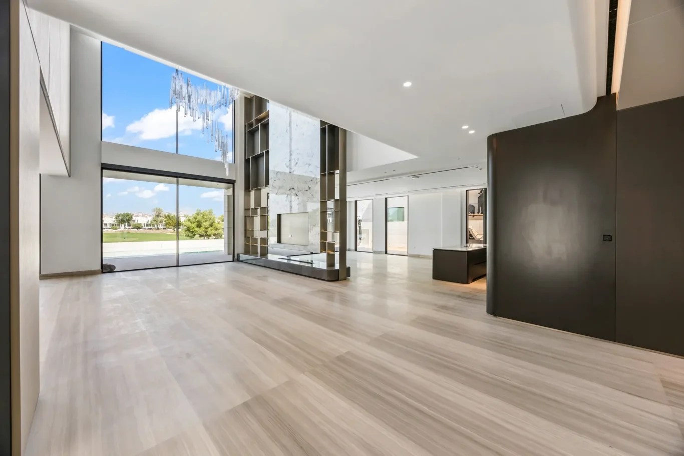 Bright open living area with floor-to-ceiling windows, marble accent wall, and a modern chandelier above.”,