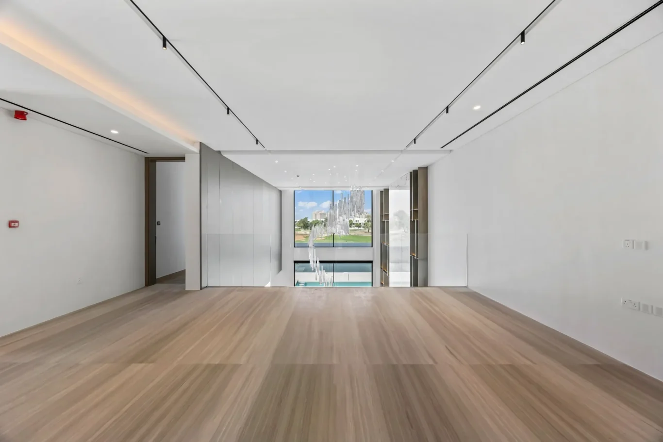Bright modern office atrium with a long open hallway, glass walls, and a multi-tier chandelier overlooking a courtyard visible through a large window.