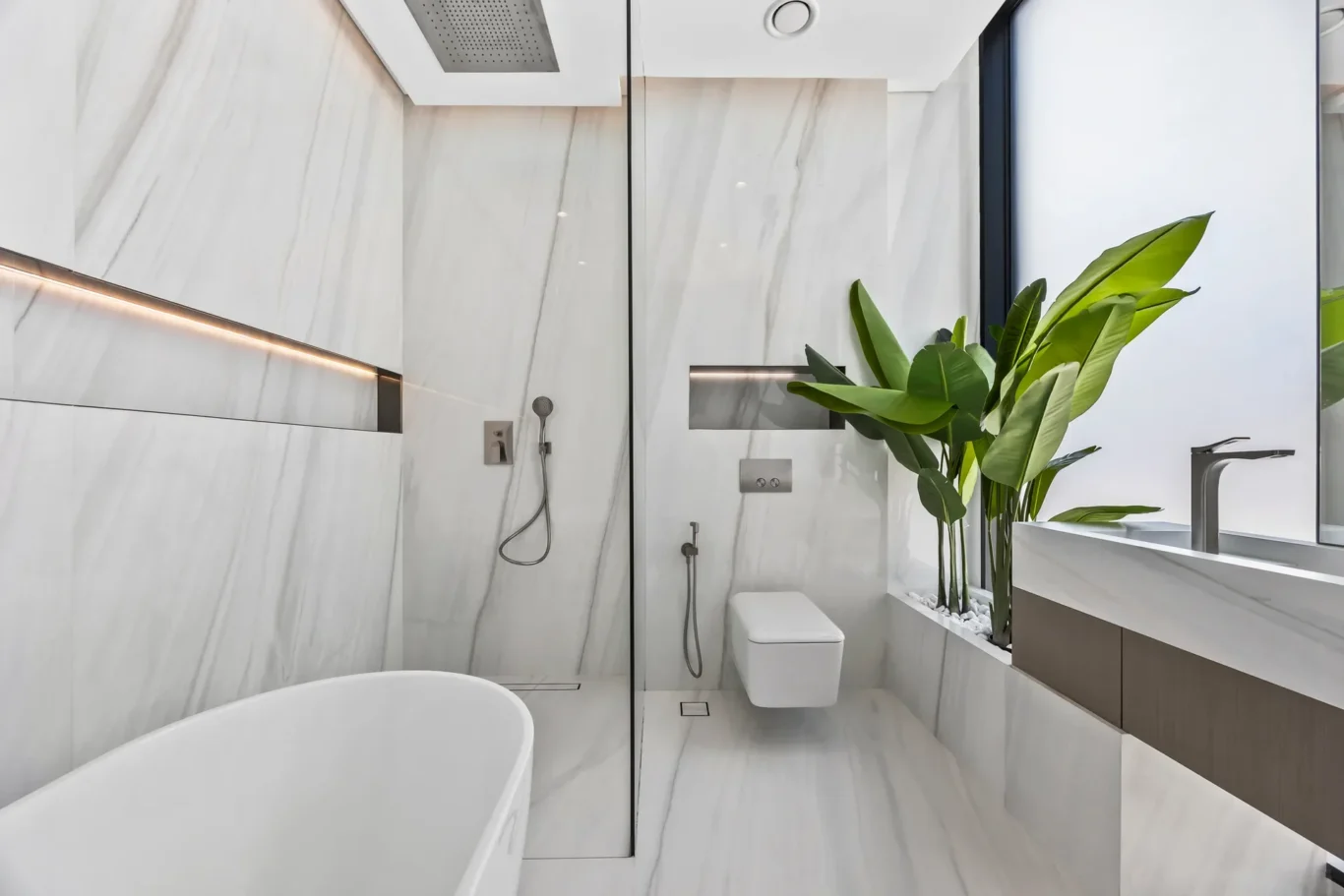 Modern luxury bathroom with marble walls, a glass shower enclosure, wall fixtures, and a large green plant by the sink.