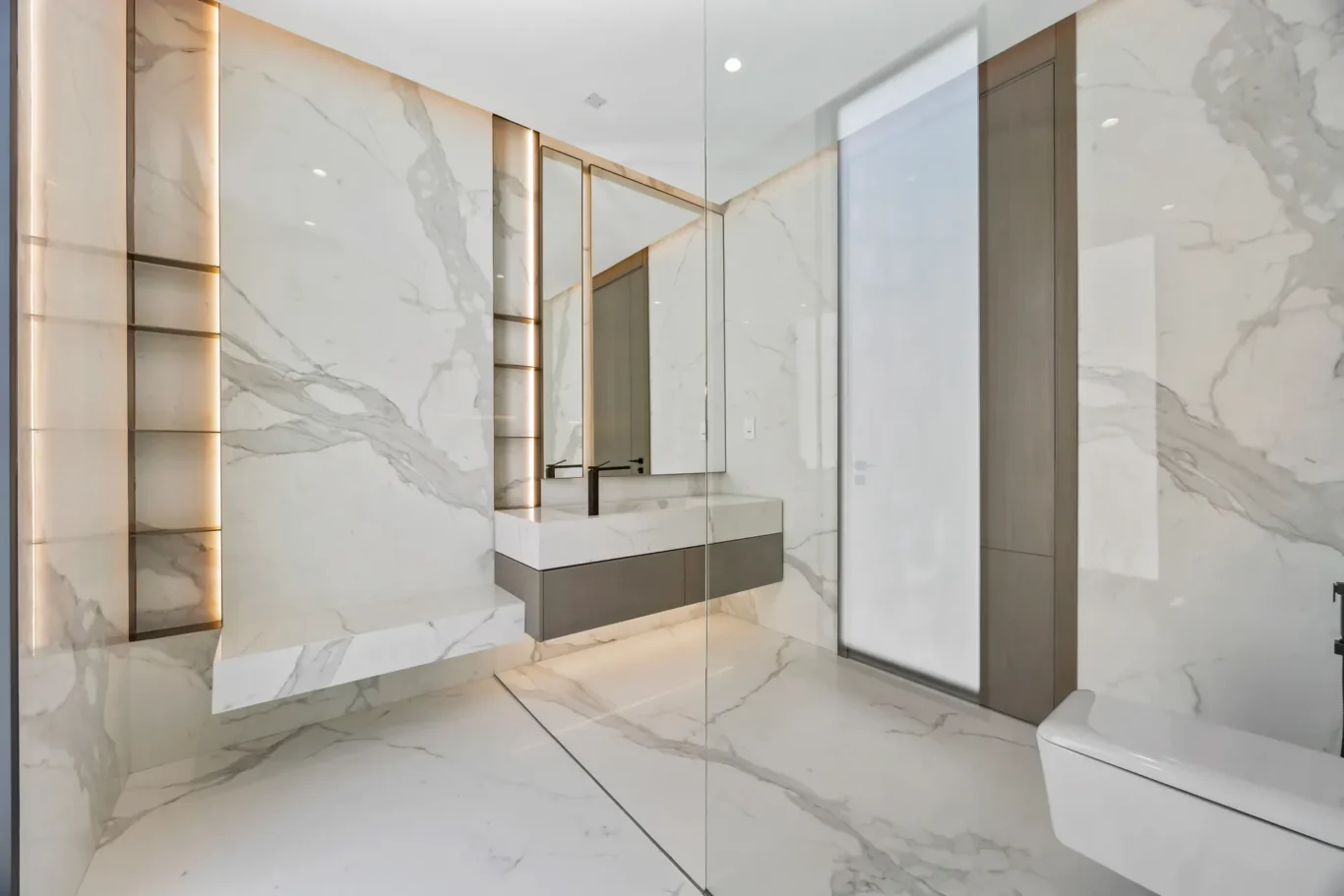 Luxurious marble bathroom with a glass-enclosed shower, wide mirror, and a floating vanity with a dark base and white countertop.