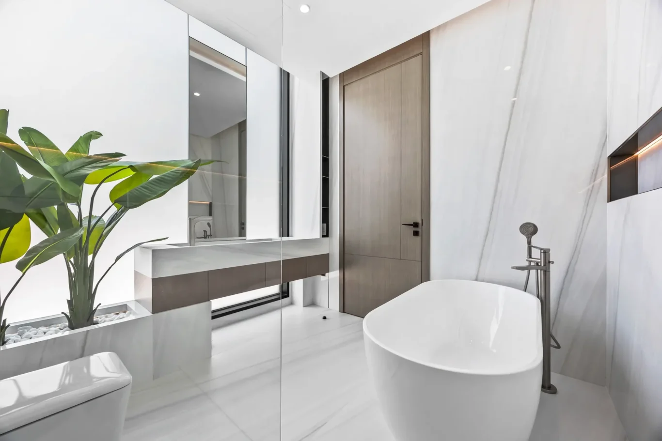 Modern bathroom with a freestanding white oval tub, glass divider, and marble walls; large green plant adds contrast.