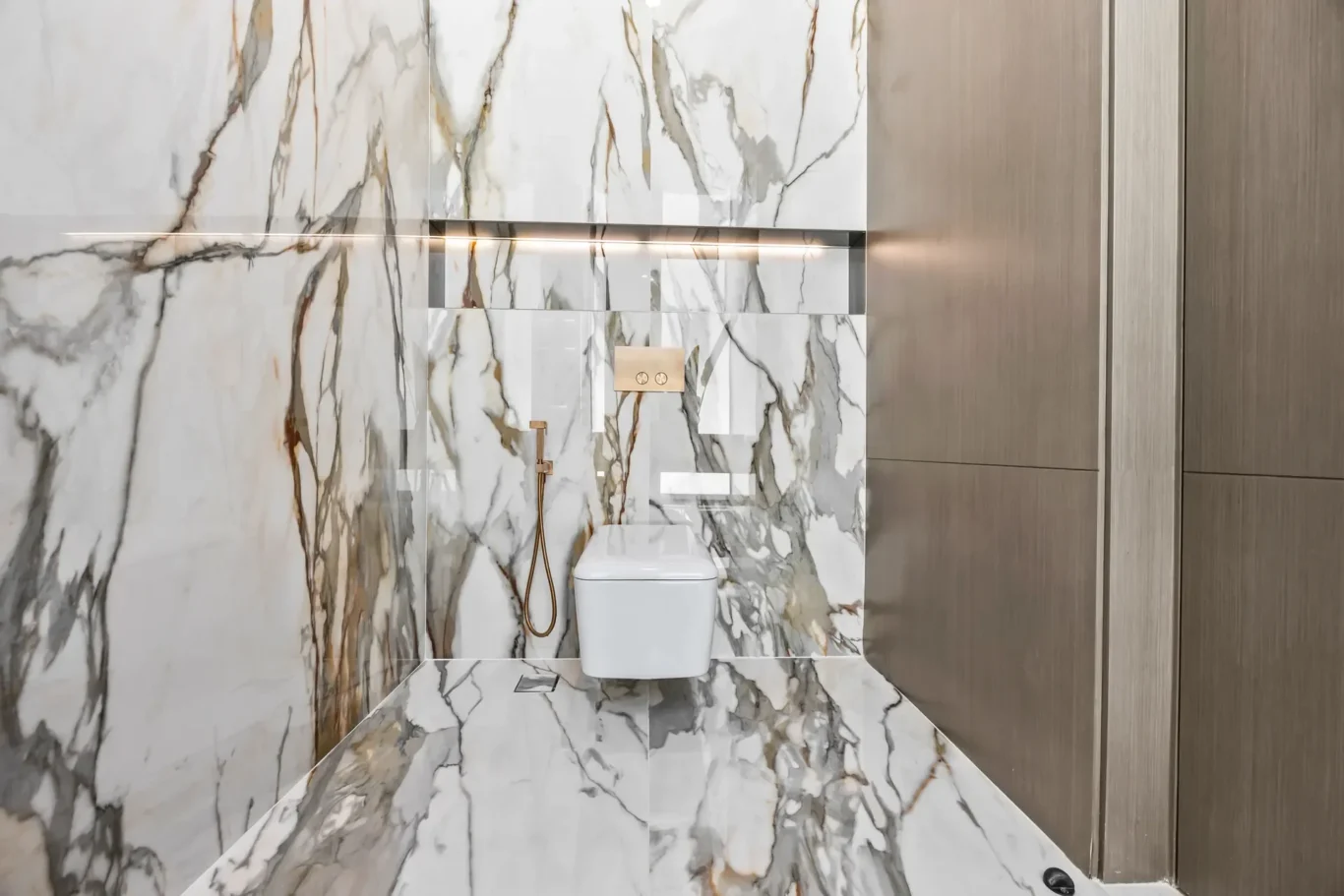 Modern bathroom with marble walls and floor, a wall-mounted toilet, and warm wood-paneled right wall with gold fixtures.
