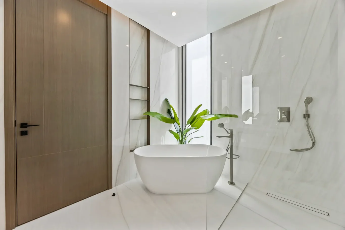 Modern minimalist bathroom with a white freestanding tub, marble walls, and a glass shower enclosure, plus a wooden door and green plant.