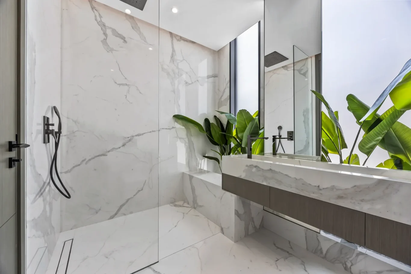 Luxurious marble bathroom with a glass shower, double sink, and tropical plants by the window.