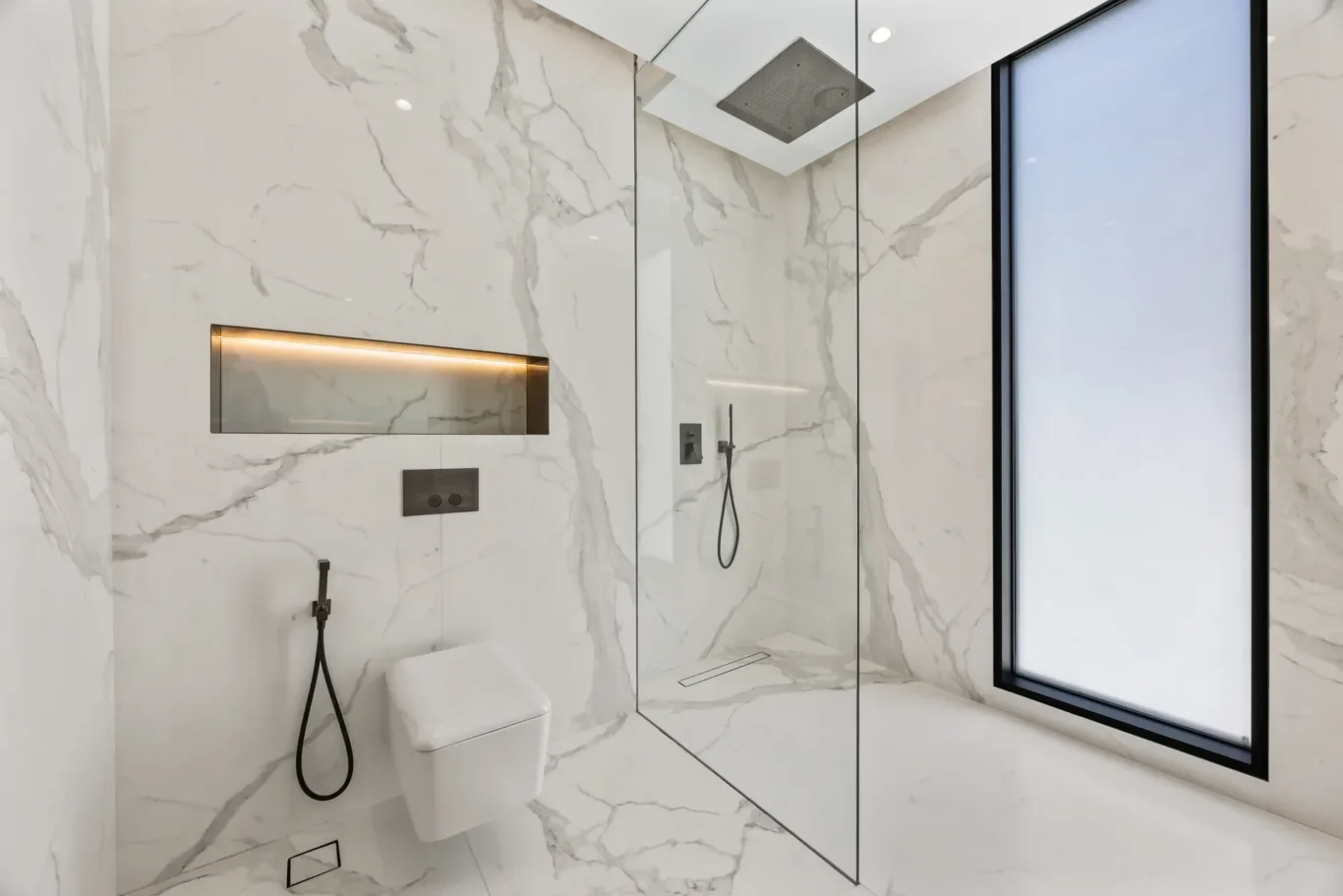 Modern marble bathroom with a glass walk-in shower, black fixtures, wall niche with warm lighting, wall-mounted controls, a white toilet, and a large frosted window.