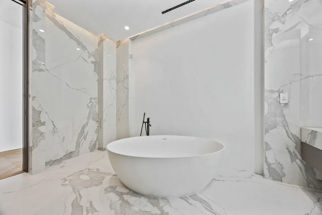 White freestanding oval tub in a marble bathroom with gray veining and a glass shower enclosure behind it.