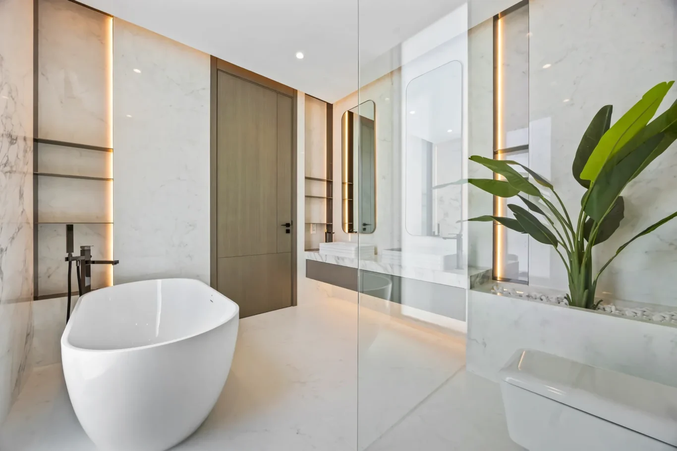 Spa-like bathroom with a freestanding white tub, marble walls, and a glass partition reflecting warm vertical lighting.