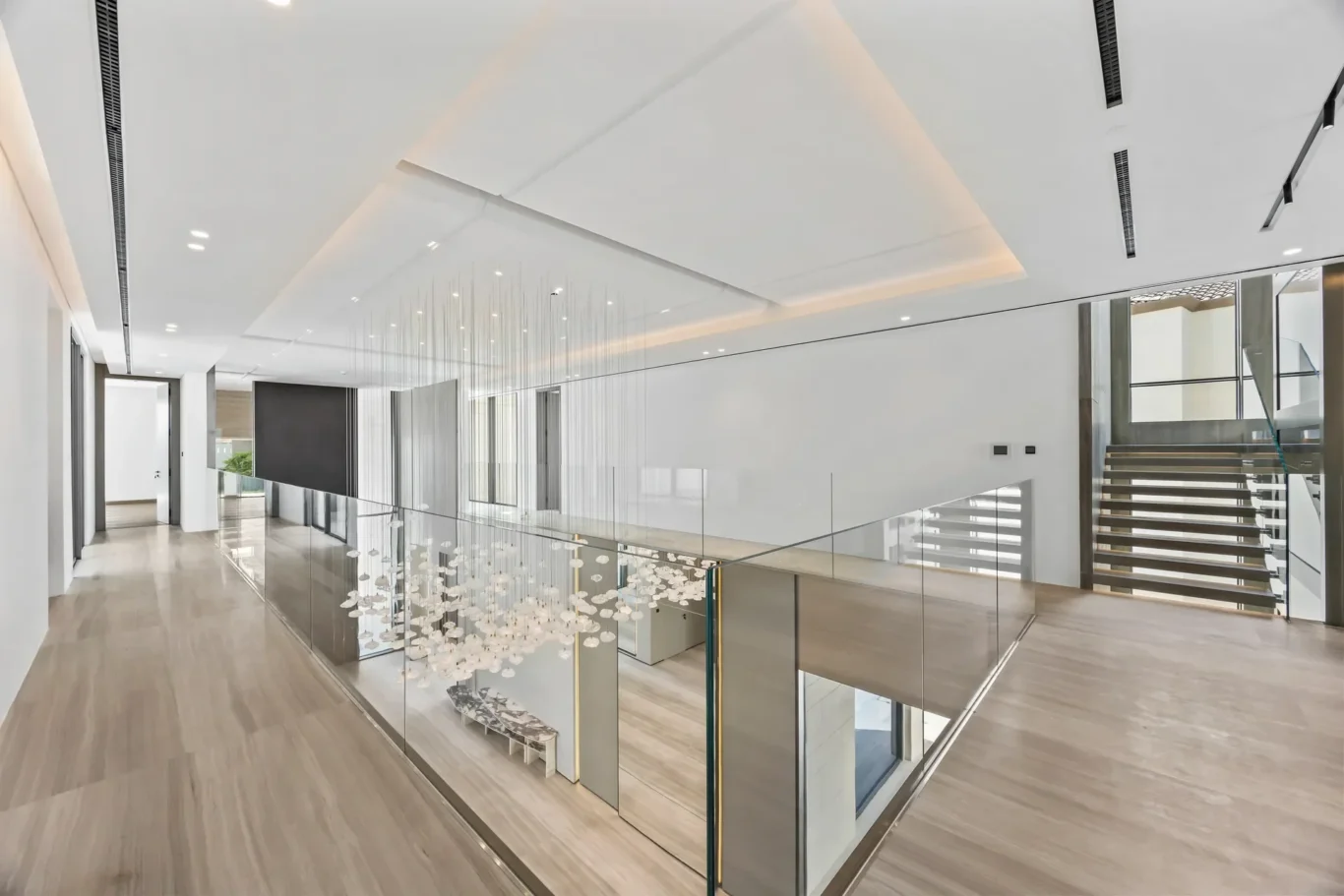 Bright modern interior: open atrium with glass railing, wooden floors, and a hanging light sculpture over a stairwell on the right side.