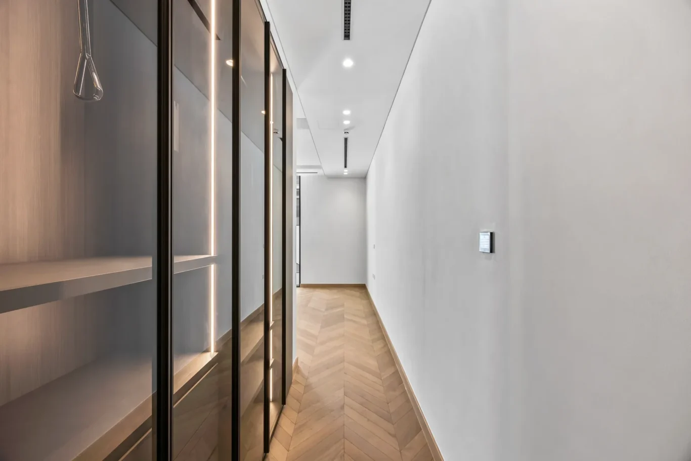Bright modern hallway with glass-fronted built-in cabinets along the left wall and a white smooth wall on the right; chevron wood floor underfoot.