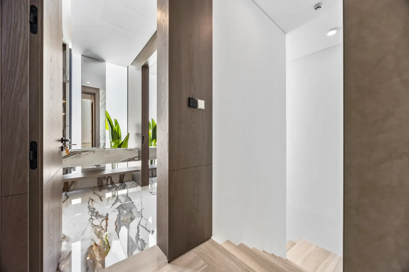 Modern hallway with marble floor, dark wood panels, and a large mirror reflecting green plants; white walls and cabinetry visible in the background.