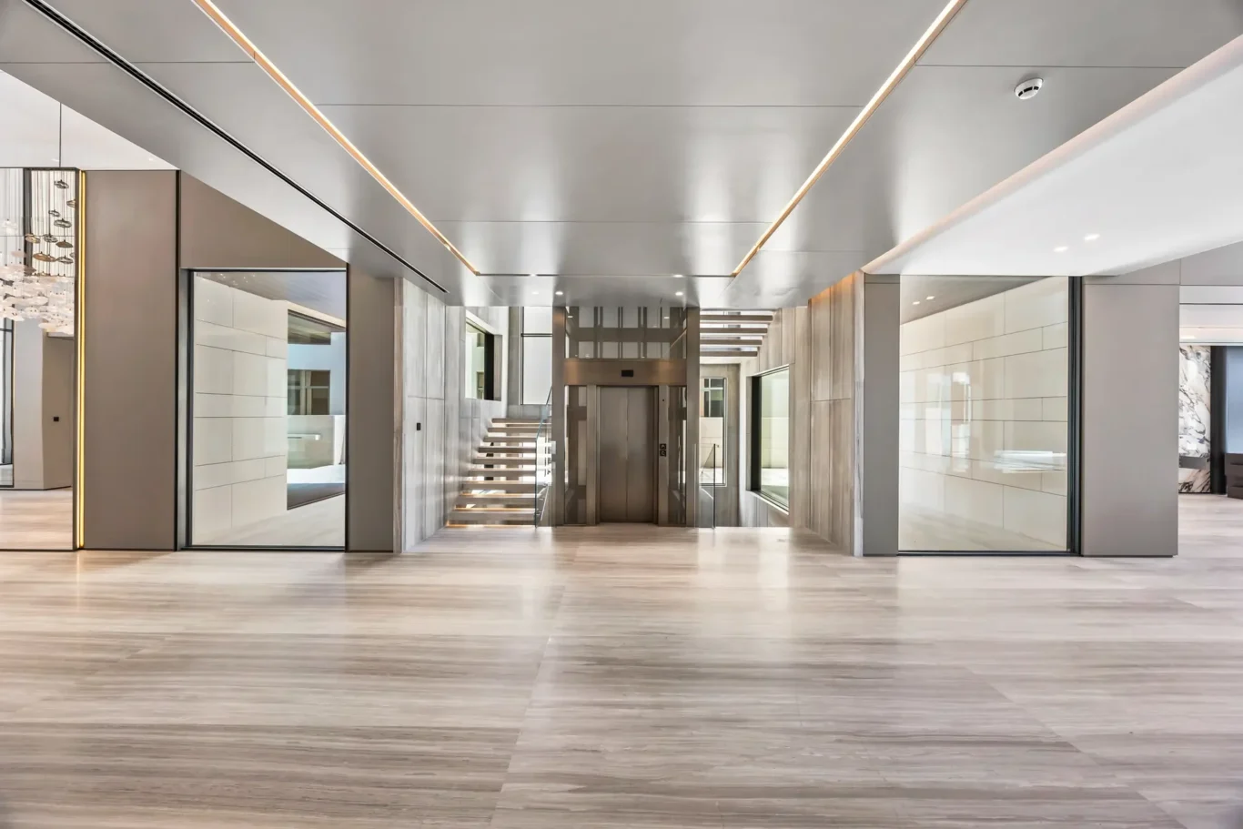 Modern hotel or office lobby with a central elevator, glass walls, and a floating staircase on the left; sleek metallic and glass surfaces with warm lighting accents.