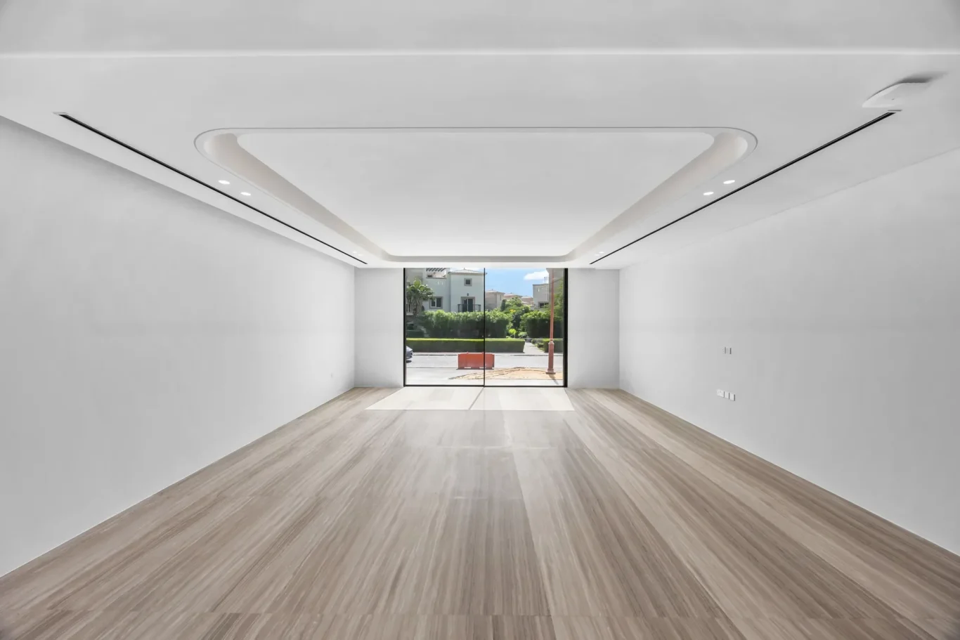 Empty modern showroom with white walls, light wood floor, and a large glass door opening to a sunny street outside.