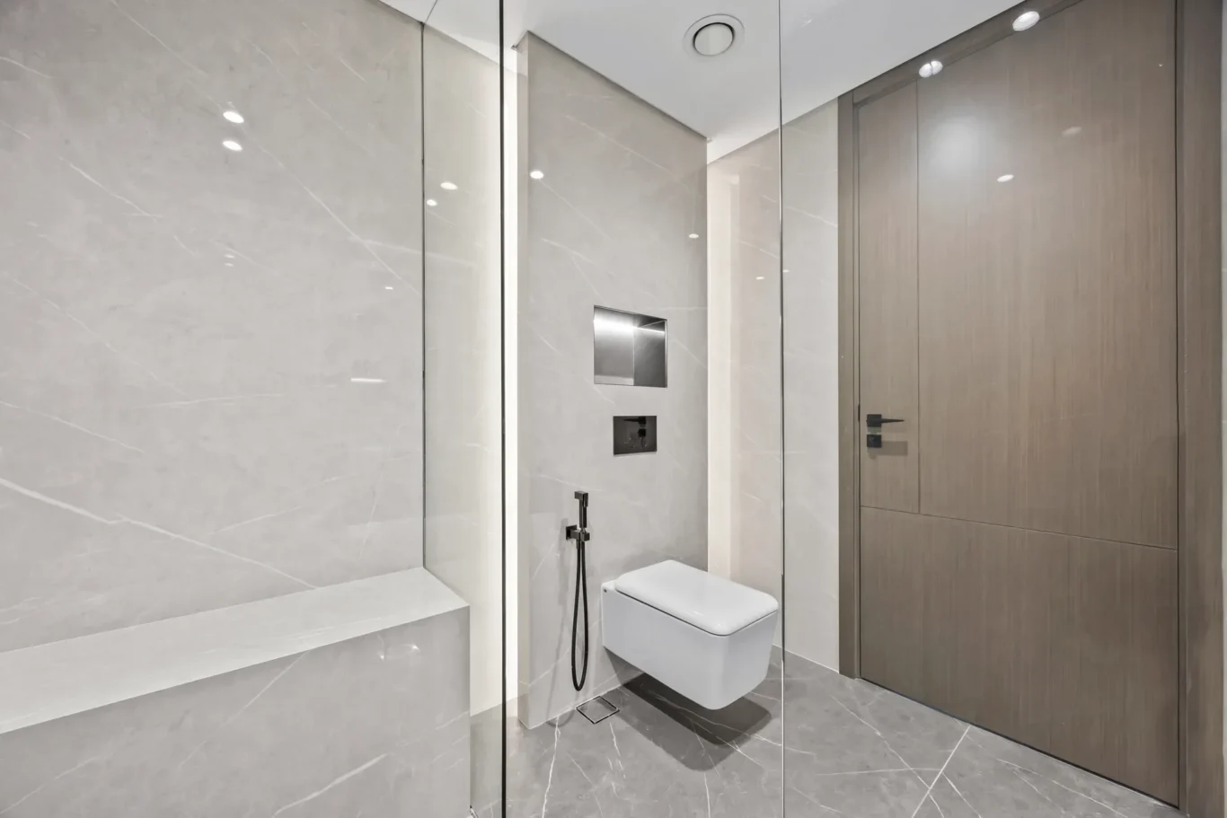 Modern bathroom with a glass shower enclosure, wall-mounted toilet, and sleek marble walls and floor; wood-toned storage cabinet on the right.