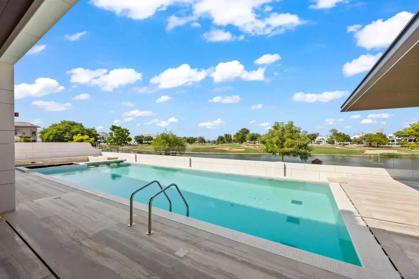 Modern outdoor pool with a wooden deck, clear blue water, and a lake view beyond the white barrier on a sunny day, with trees and houses in the distance.
