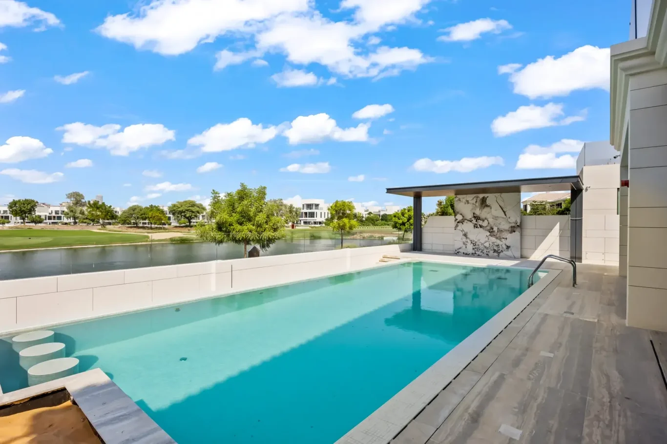 Modern poolside terrace with a turquoise rectangular pool, gray wooden deck, and a marble accent wall overlooking a lake and green trees under a blue sky.