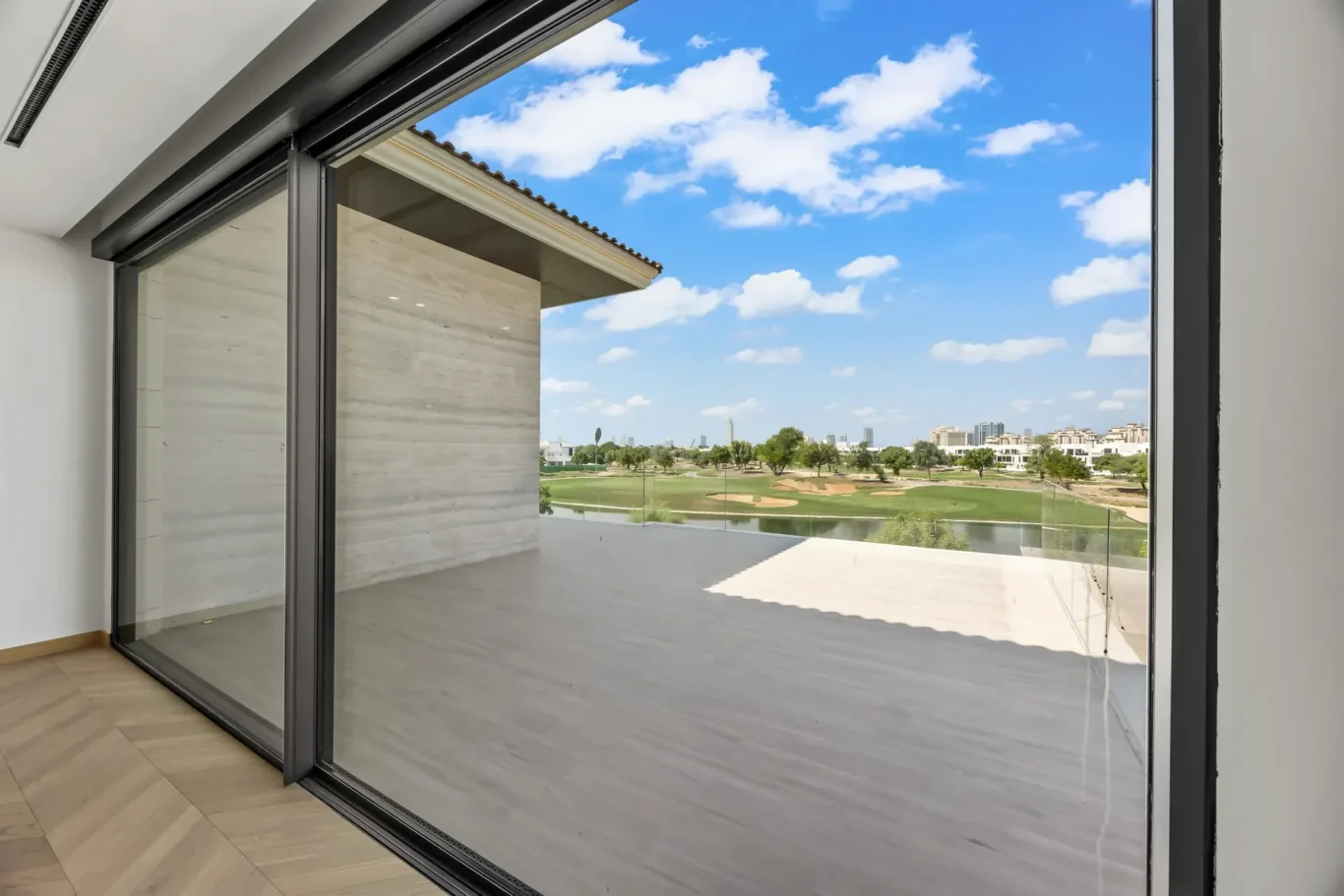 Floor-to-ceiling sliding glass doors open to a balcony with a view of a golf course and distant city skyline under a blue sky.