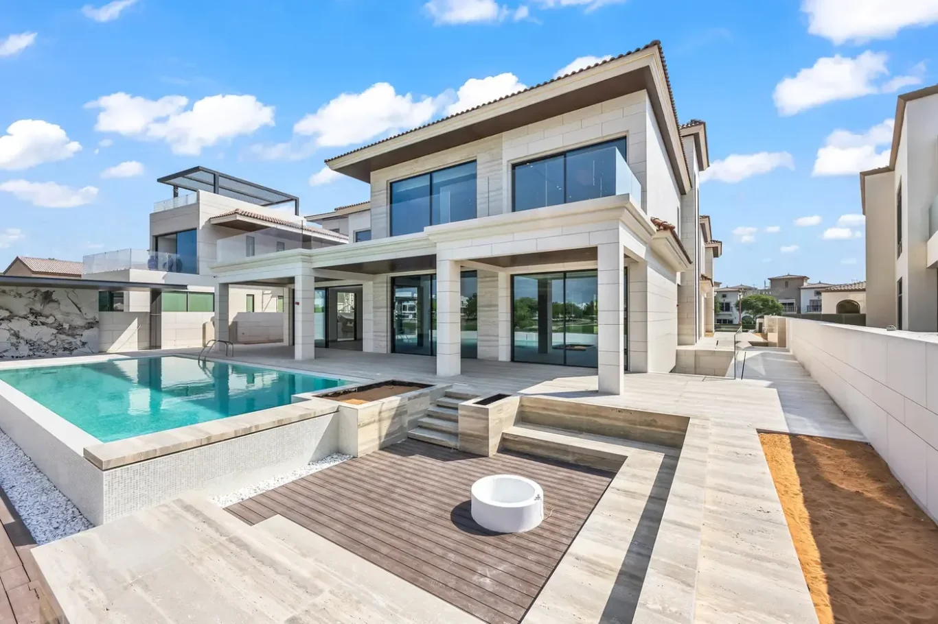 Modern two-story luxury home with a rectangular pool and spacious beige stone patio under a blue sky.