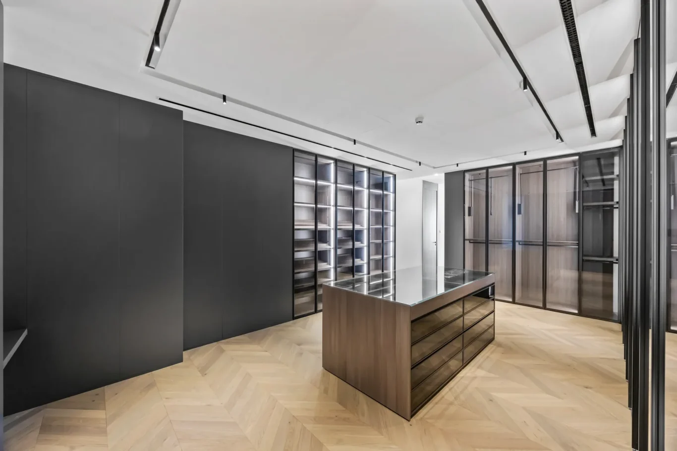 Modern walk-in closet with dark matte cabinets, glass-front wardrobes, and a central wooden island with a glass top on a herringbone wood floor