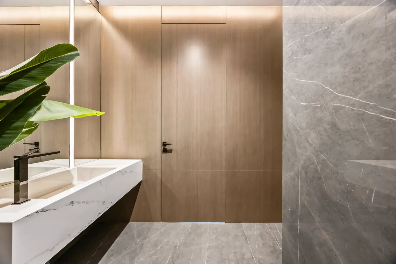 Modern bathroom with gray marble walls, white marble sink countertop, black faucet, and warm wood cabinetry with a green plant nearby.