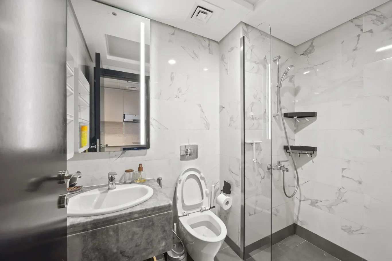 Modern marble bathroom with glass shower enclosure, white toilet, sink on a gray marble countertop, and a large mirror above.