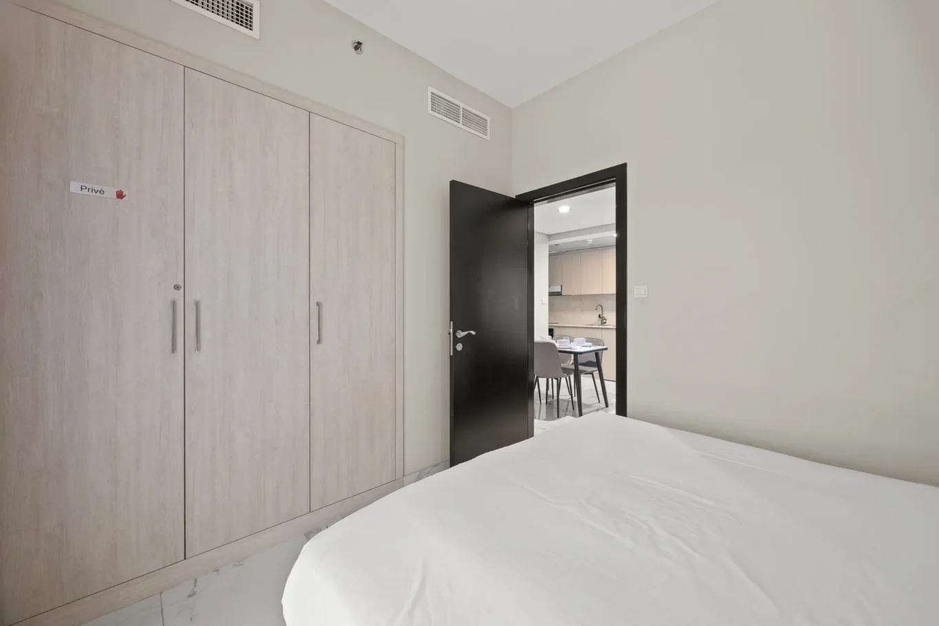 Bedroom with light wood built-in wardrobes along the left wall, white bed in the foreground, and an open doorway to a kitchen/dining area.
