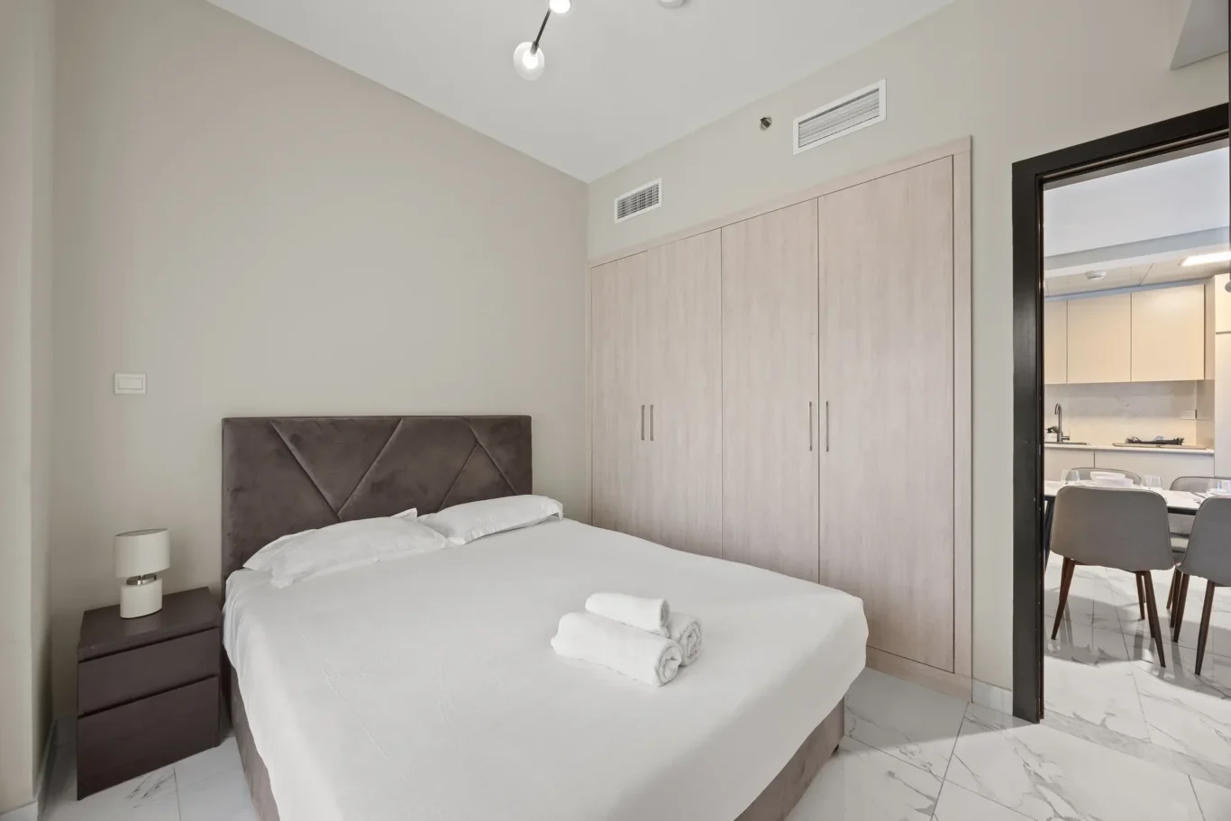 Modern bedroom with white bedding, grey upholstered headboard, dark nightstand, and pale wood built-in wardrobes; doorway to a kitchen on the right.