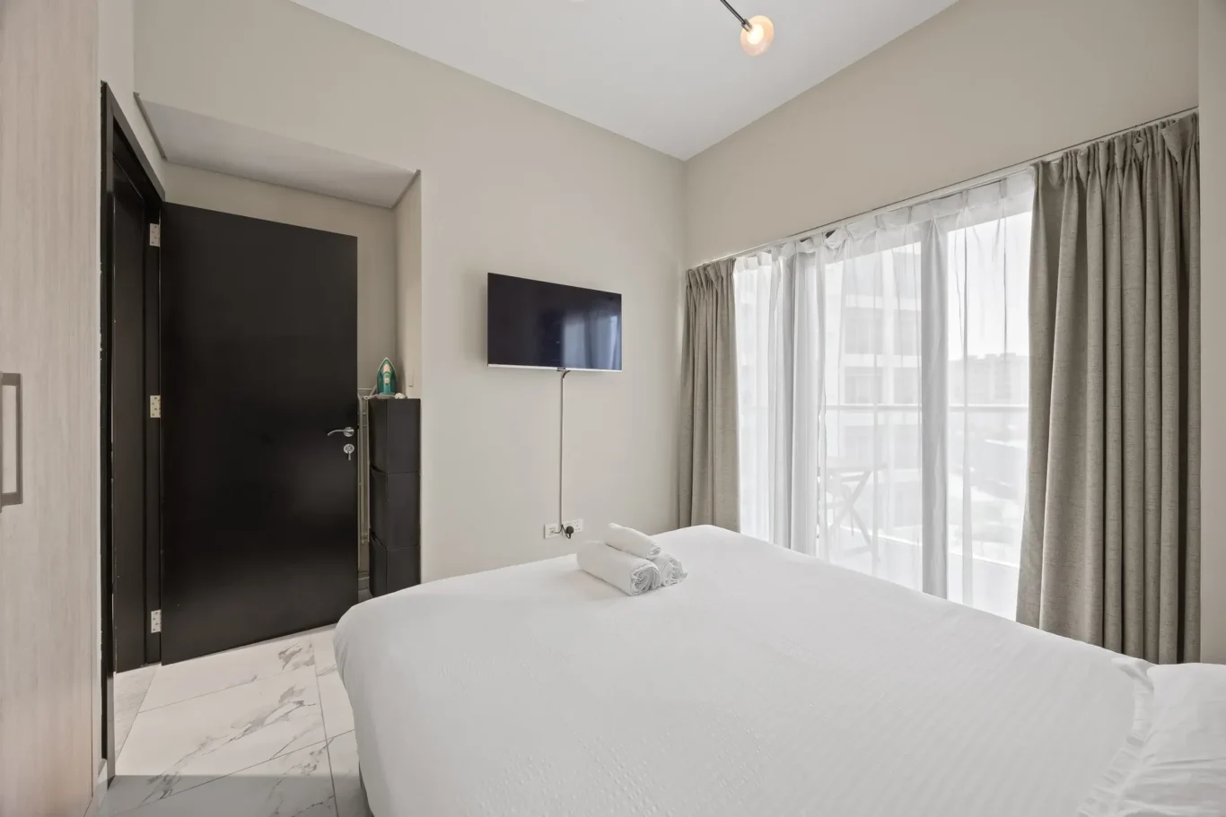 Hotel room with two white beds, rolled towels on the near bed, a wall-mounted TV, and large windows with sheer gray curtains.